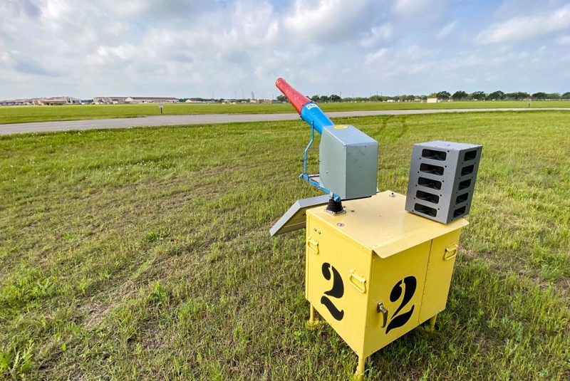 A squat yellow metal cabinet situated on a grassy area with a blue and red horn attached, pointing toward a facility in the distance.
