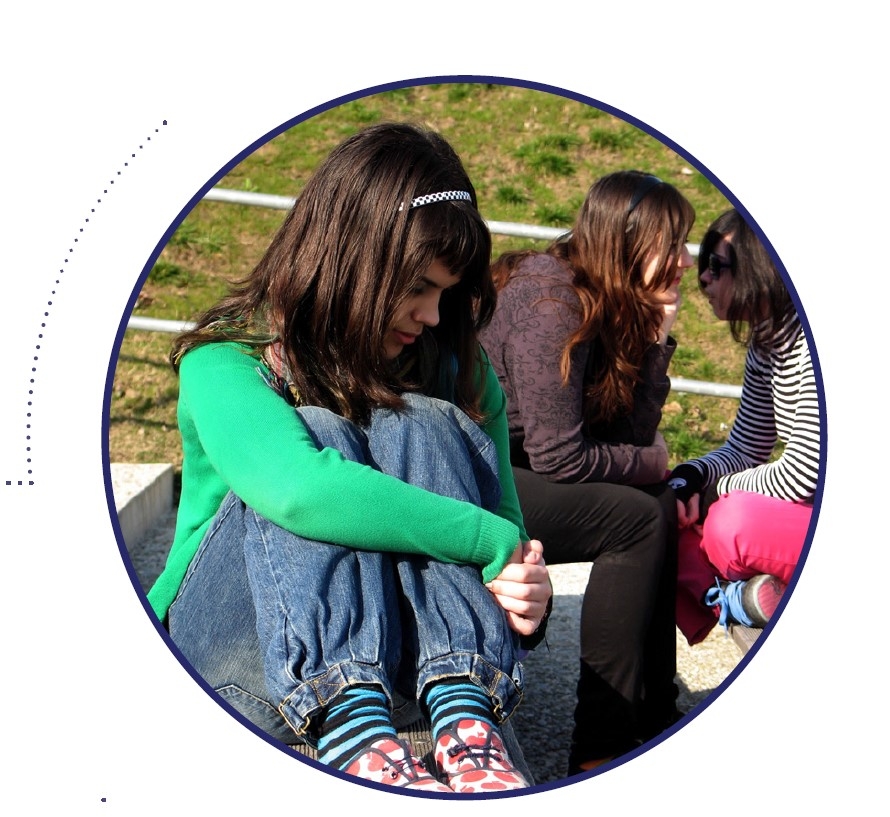 Picture of a sad girl sitting alone and two other girls in the background talking to each other.