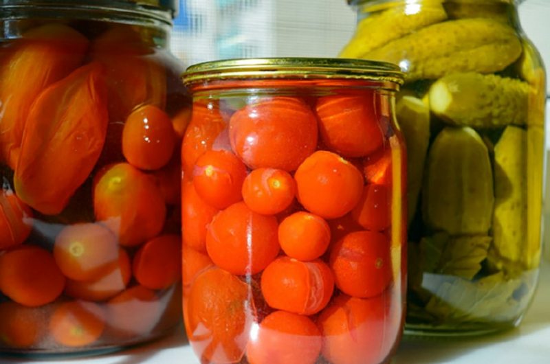 Two clear glass jars of home-canned red tomatoes next to a similar jar of green pickles.