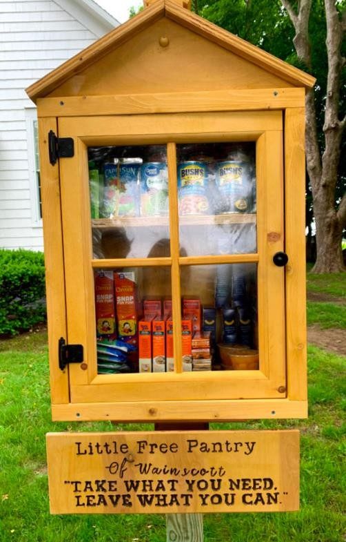 A share pantry, made of wood with a framed door on the front. There are two shelves inside, filled with nonperishable foods. There is a sign on the bottom of the pantry saying "Little Free Pantry of Wainscott. 'TAKE WHAT YOU NEED, LEAVE WHAT YOU CAN."