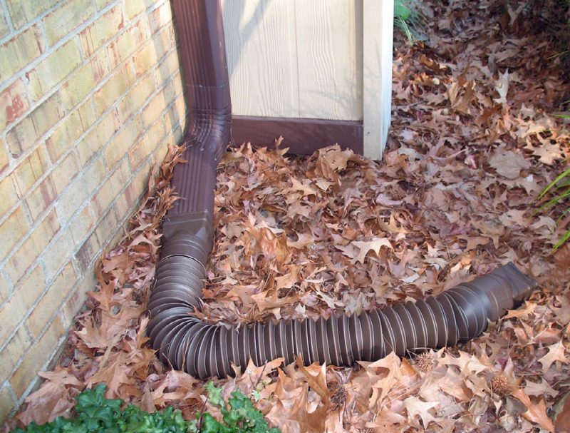 A brown extender on a brown downspout in a pile of leaves next to a brick exterior wall.