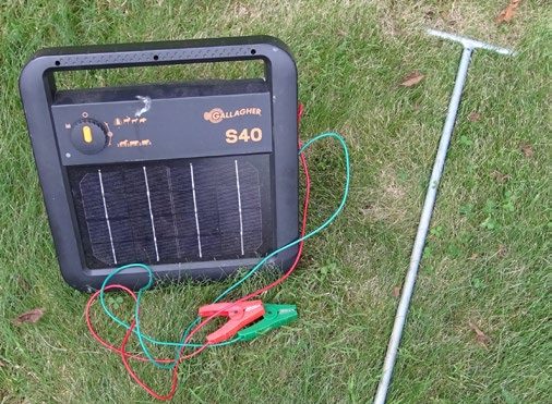 A solar energizer with red and green lead wires and clamps and a portable galvanized steel ground rod laid out in some grass.